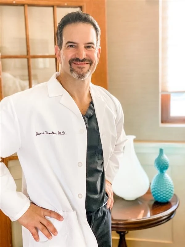 Man with dark hair and goatee wearing white coat stands with hands on hips in a room with window and table.