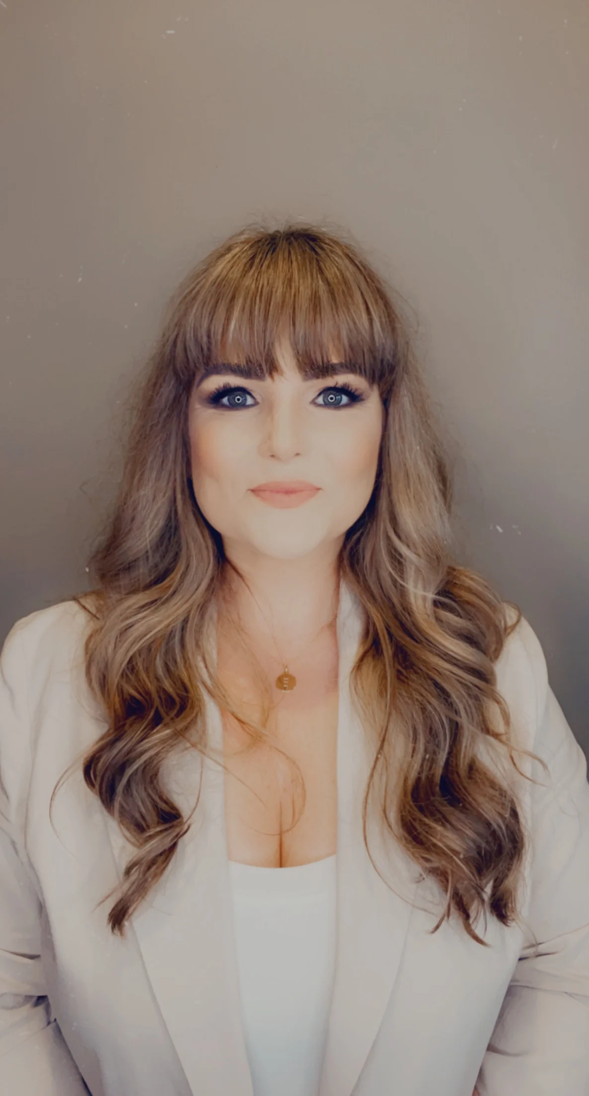 Woman with long wavy hair and bangs wearing a light blazer and necklace against a plain background.