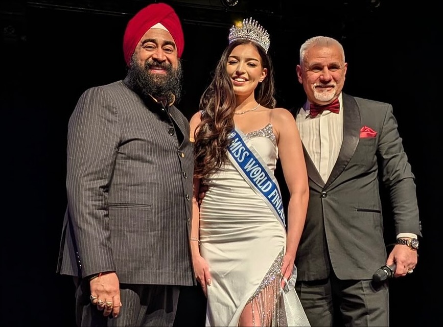 Miss World finalist wearing a crown and sash stands smiling between two men in formal suits on stage.