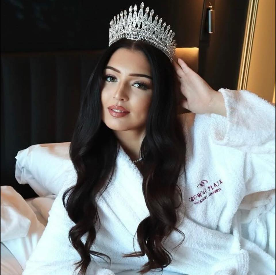 Woman with long dark hair wearing a jeweled crown and white Crown Plaza robe seated on a bed.