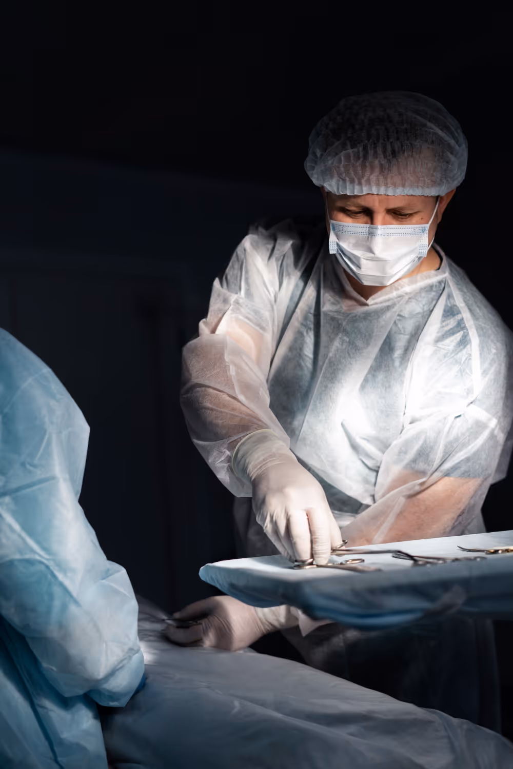 Surgeon in protective clothing and mask selecting surgical instruments on a tray in an operating room.