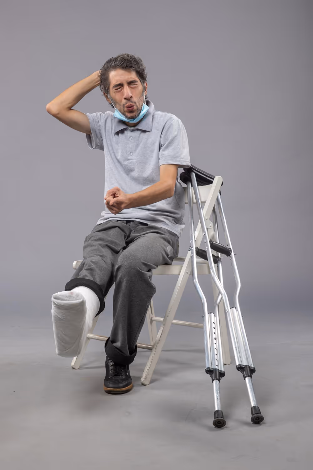 Man with a cast on his left foot sitting on a white chair with crutches beside him, making a face while scratching his head.