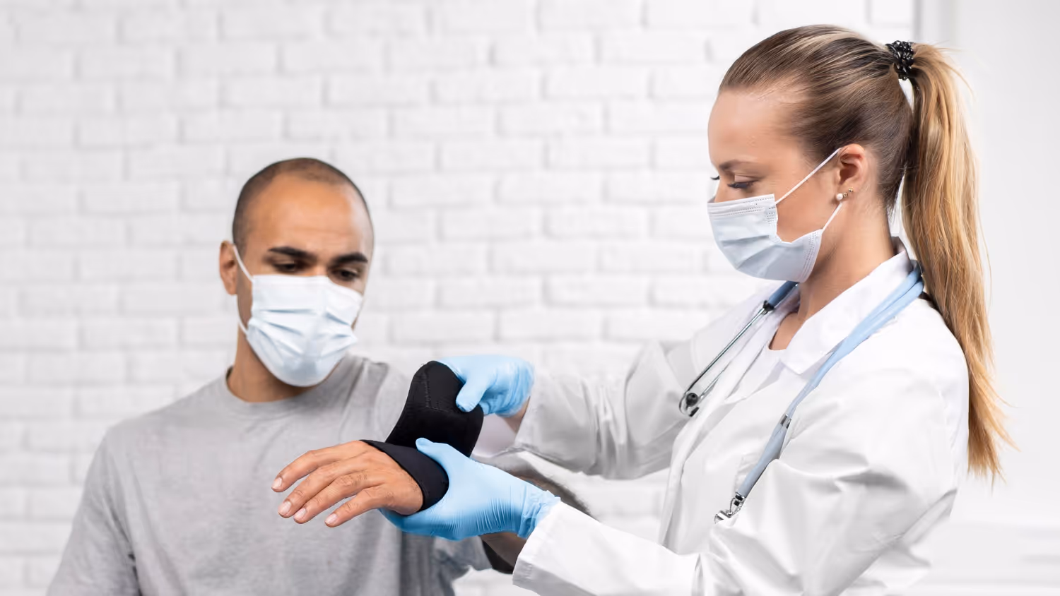 Healthcare professional wearing mask and gloves wraps a wrist brace around a male patient's wrist.