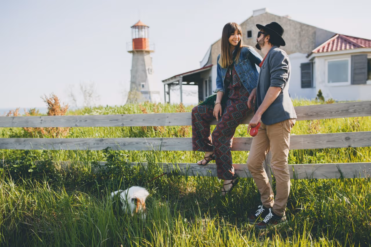 Young woman sitting on a wooden fence smiling at a man holding a dog leash, with a fluffy white dog and a lighthouse in the background.