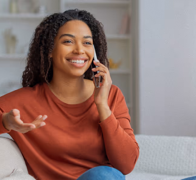 Smiling woman with curly hair wearing a rust-colored sweater talking on a smartphone indoors.
