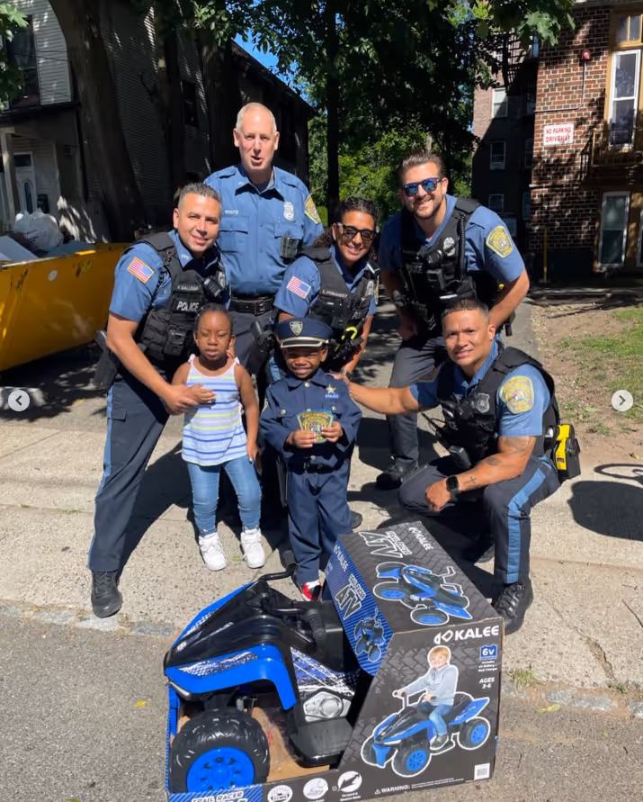 A group of police officers posing for a picture outdoors, standing on the ground with trees in the background. They are wearing uniforms and various types of footwear.


