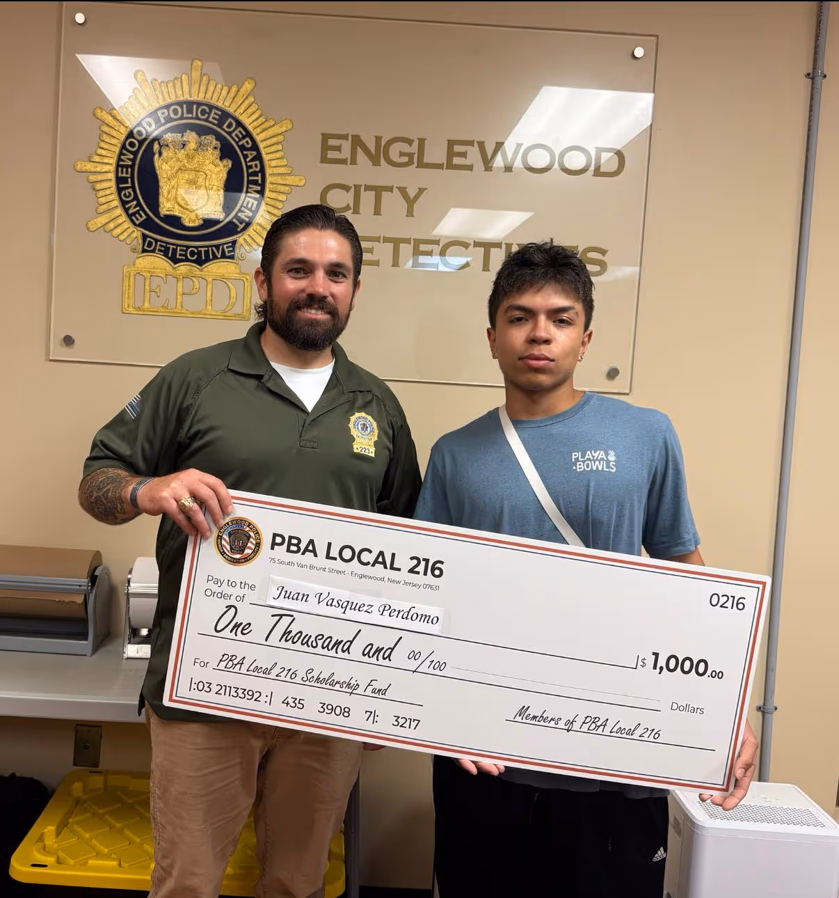 Two men are standing indoors, smiling as they hold a large check. The check has handwritten text indicating it is for "Juan Vasquez Perdomo" in the amount of one thousand dollars for the PBA Local 216 Scholarship Fund. They are dressed in casual clothing, and there is a whiteboard or wall behind them.