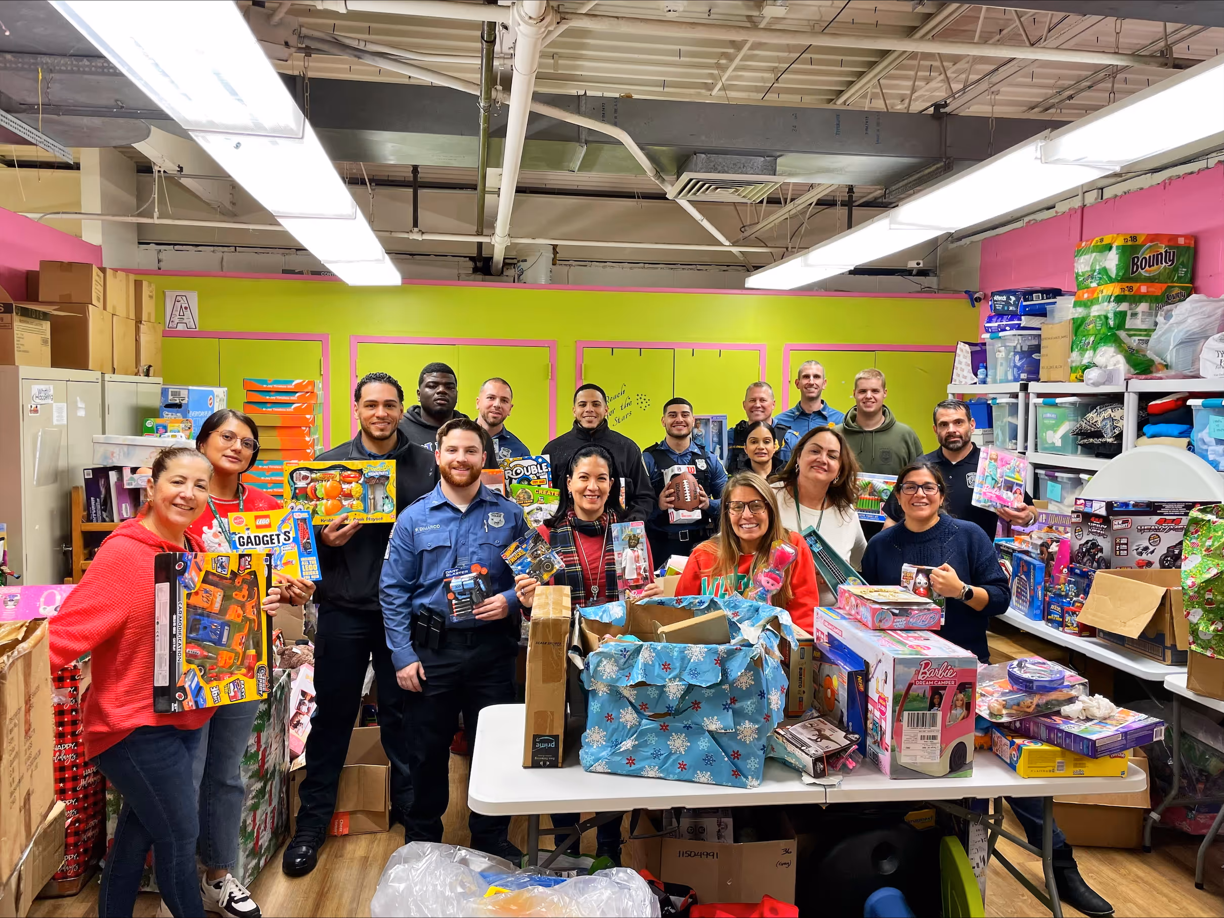 A group of people posing for a photo indoors, smiling and standing together in a shop with visible clothing and footwear. Various individuals are gathered near a table against a wall, with the ceiling and floor visible in the room.


