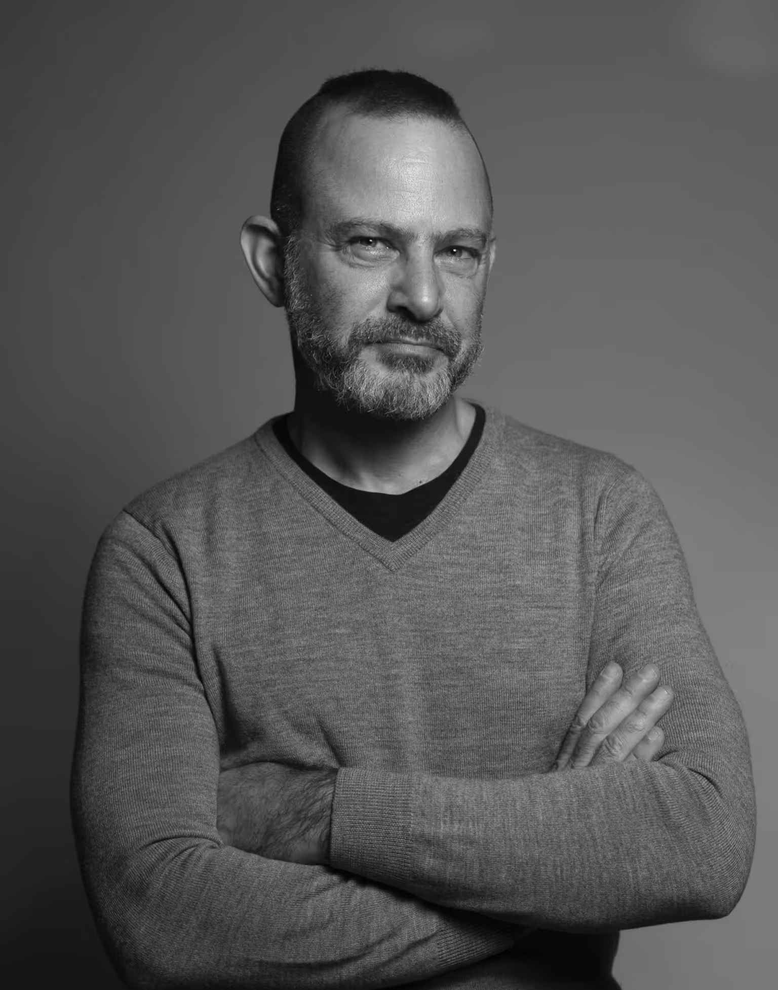 Black and white portrait of a middle-aged man with a beard and short hair, wearing a V-neck sweater, arms crossed, and looking confidently at the camera.