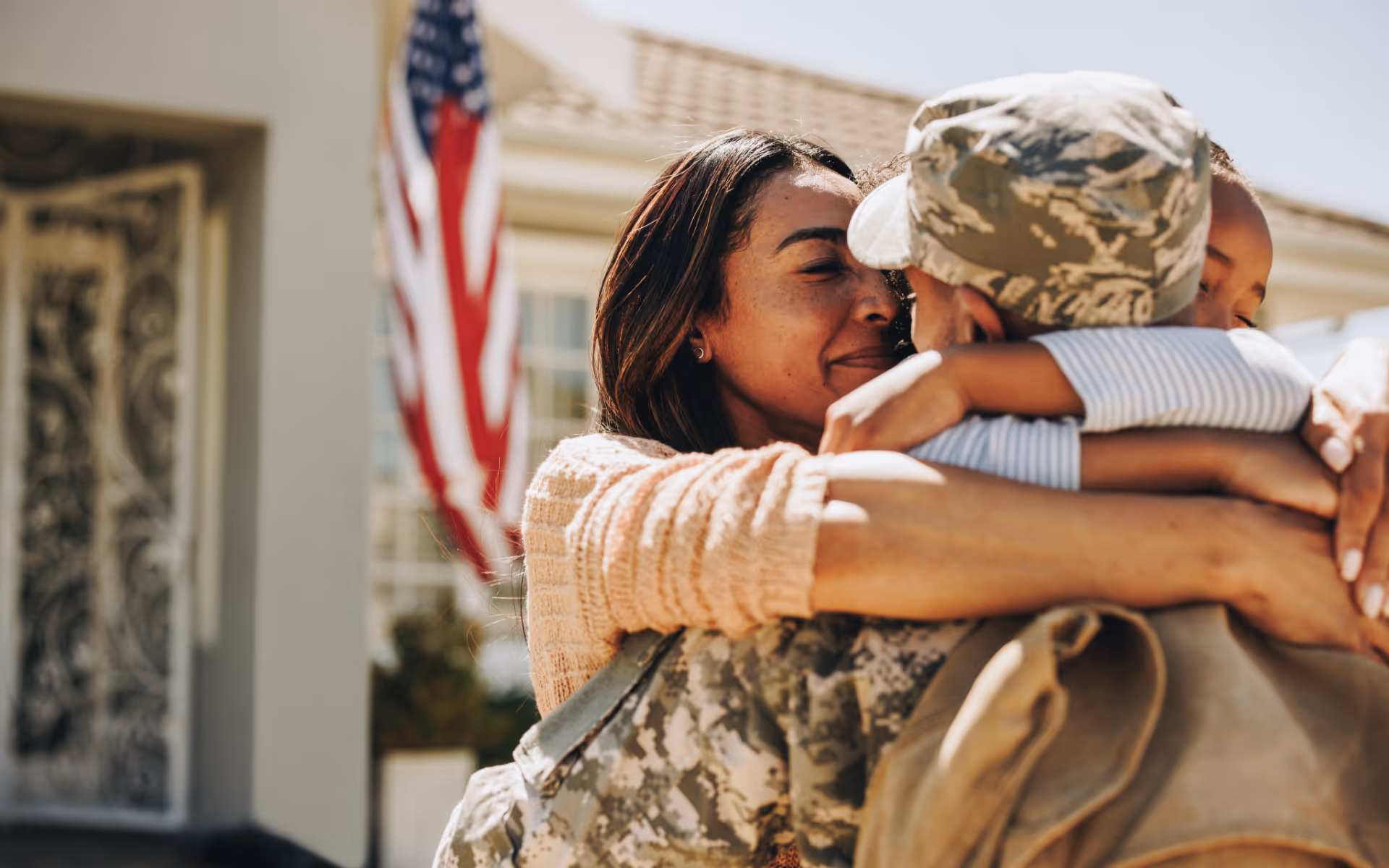 Woman and child warmly hugging a soldier in camouflage uniform in front of a house with an American flag.