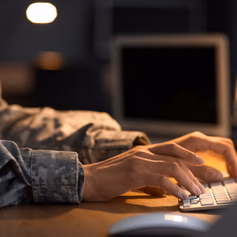 Person in military uniform typing on a keyboard at a desk with a computer monitor in the background.
