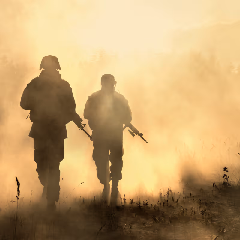 Silhouettes of two armed soldiers walking through a foggy or smoky field during sunset or sunrise.