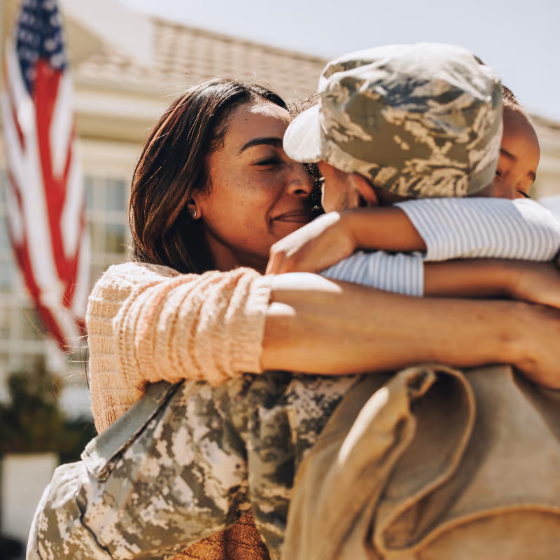 Woman hugging a soldier in camouflage uniform outside with an American flag in the background.