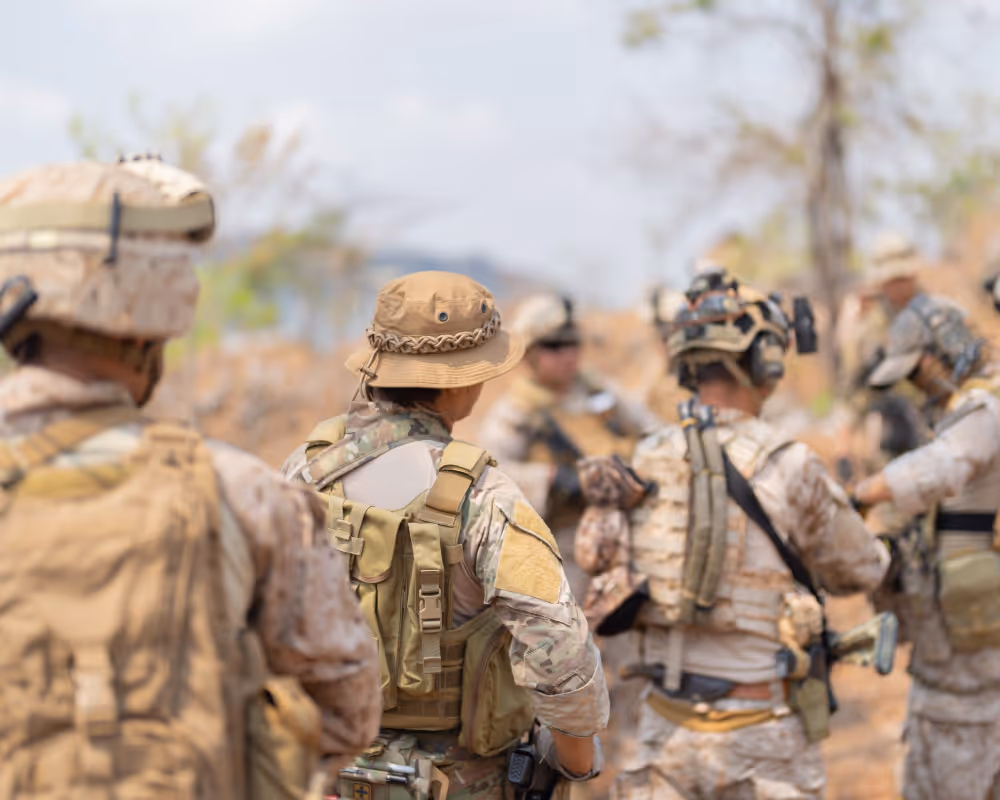 Group of soldiers in camouflage gear and tactical vests standing outdoors in a dry, open area.