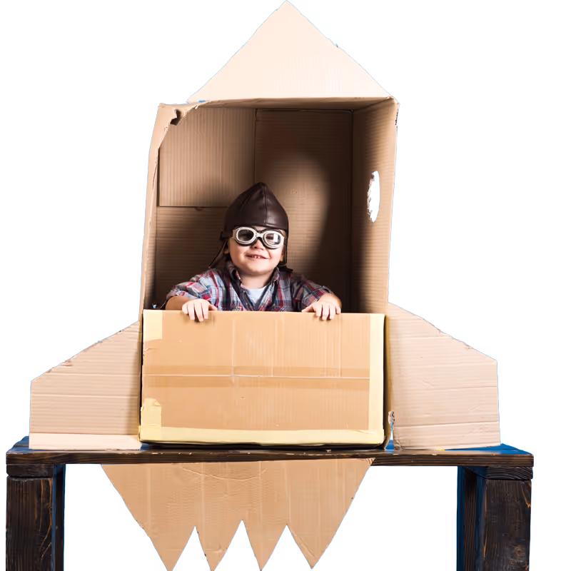A smiling child wearing aviator goggles and a leather pilot hat inside a cardboard rocket ship against a blue background.