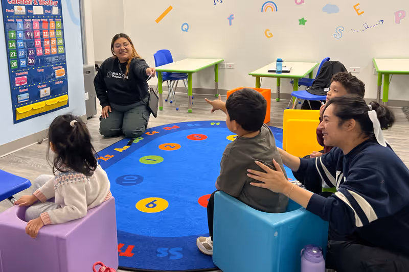 Teacher kneeling on a colorful number rug, engaging with three children seated in small chairs in a classroom.