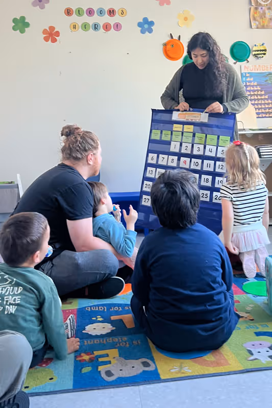 Teacher standing in a classroom showing a large calendar chart to a group of young children sitting on a colorful alphabet mat.
