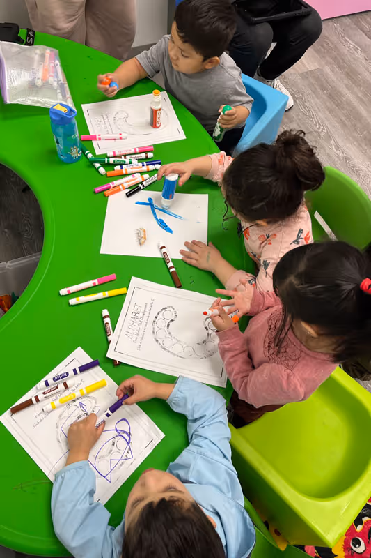 Four young children sitting around a green table, coloring alphabet worksheets with markers and glue sticks.