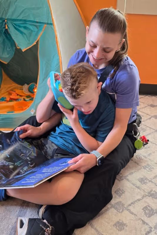 Woman sitting on floor hugging a child who is holding a tablet and looking at a book together.