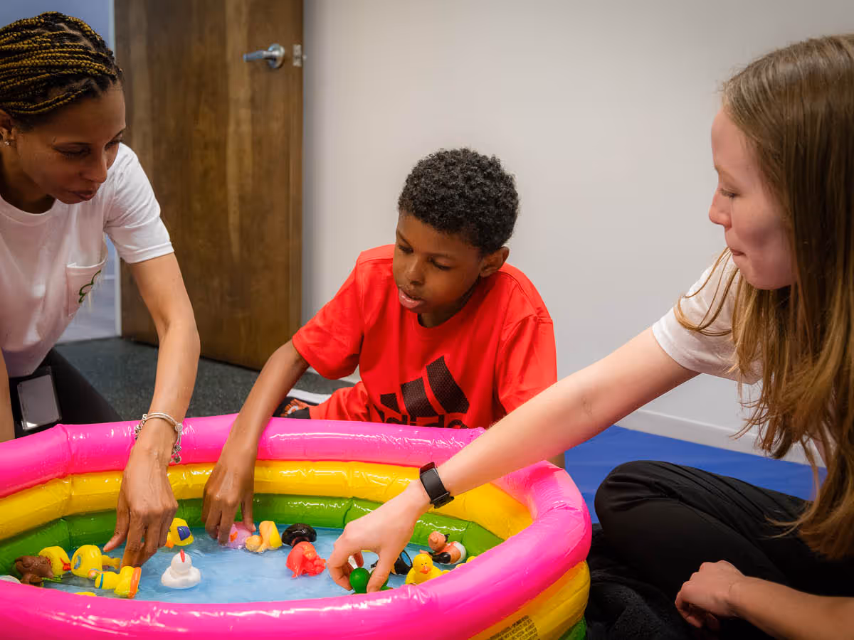 Two women and a boy playing with floating toys in a small colorful inflatable pool indoors.