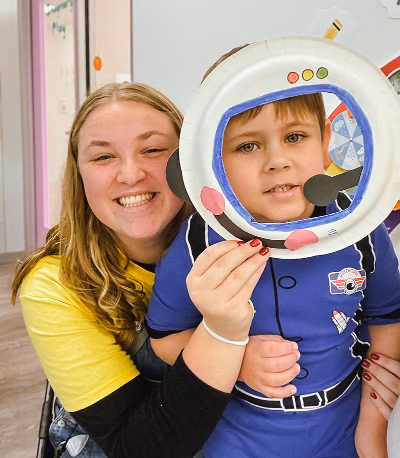 Smiling woman with blonde hair hugging a young boy who is holding a paper astronaut helmet cutout in front of his face.