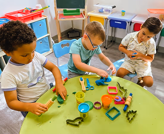 Three children sitting around a green table playing with colorful modeling clay and sculpting tools in a classroom.