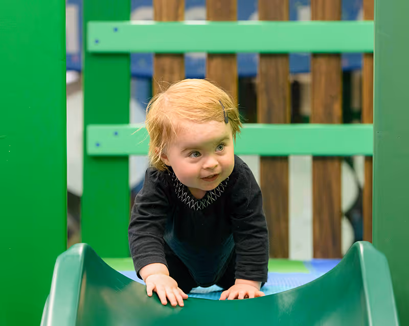 Young child with blonde hair and a hair clip crawling on a green slide in a playground.