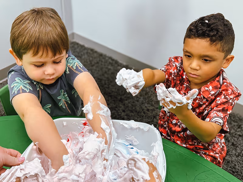 Two young boys playing with foam or shaving cream in a large white container on a green table.