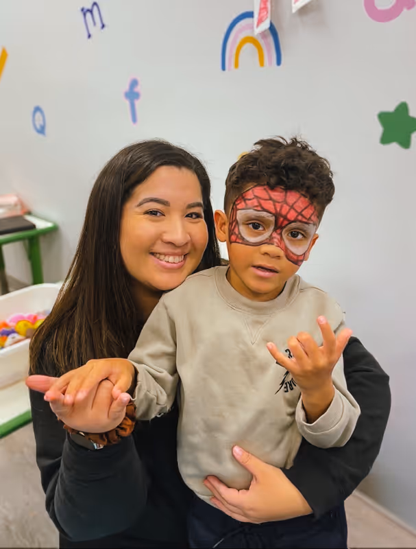 Smiling woman hugging a young boy with Spiderman face paint in a colorful playroom.