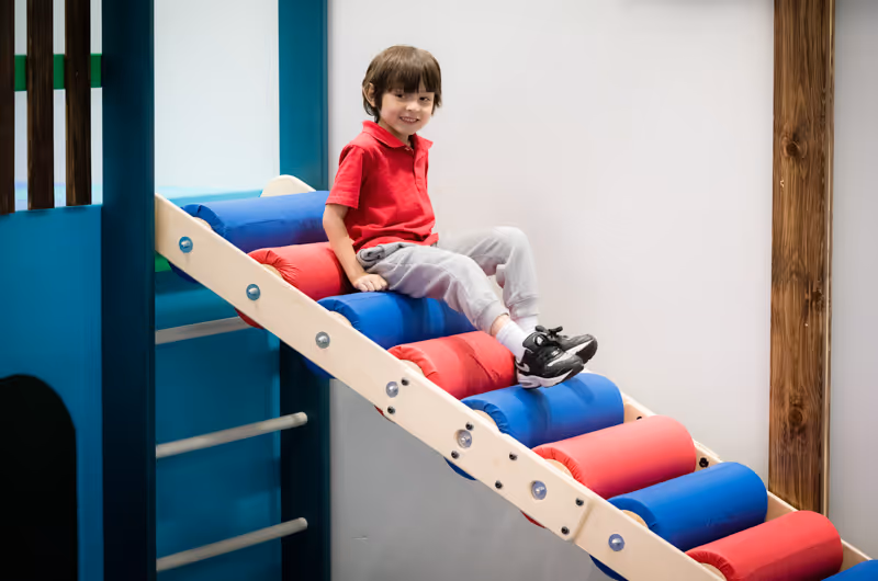 Child in red shirt and gray pants sitting and smiling on a colorful indoor play structure with blue and red padded steps.