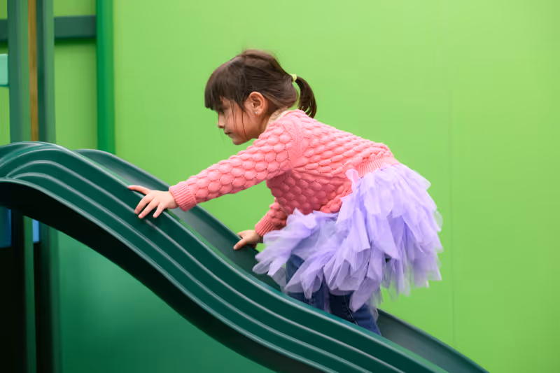 Young girl wearing a pink sweater and purple tutu climbing up a green slide indoors.