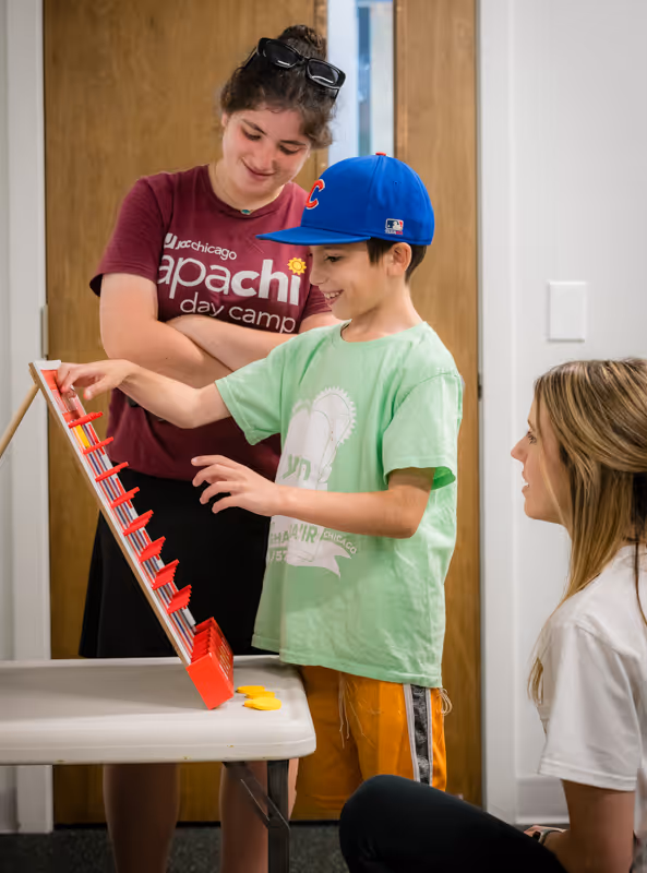 A boy in a blue baseball cap and green shirt plays a pegboard game on a table while two women watch and smile.