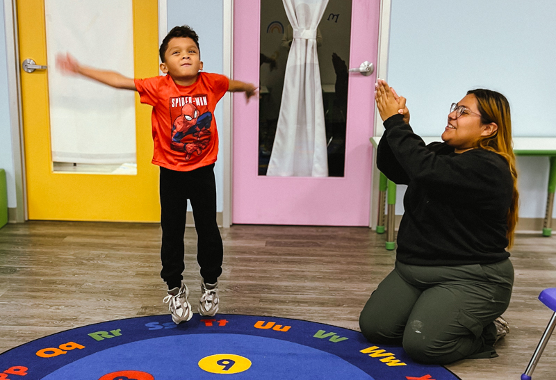 A young boy wearing a red Spider-Man shirt jumps on a colorful circular alphabet rug while a woman kneels nearby clapping and smiling.