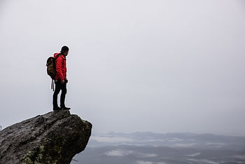 Man in red jacket with backpack stands on a cliff edge, overlooking misty mountains. Overcast sky, contemplative mood.