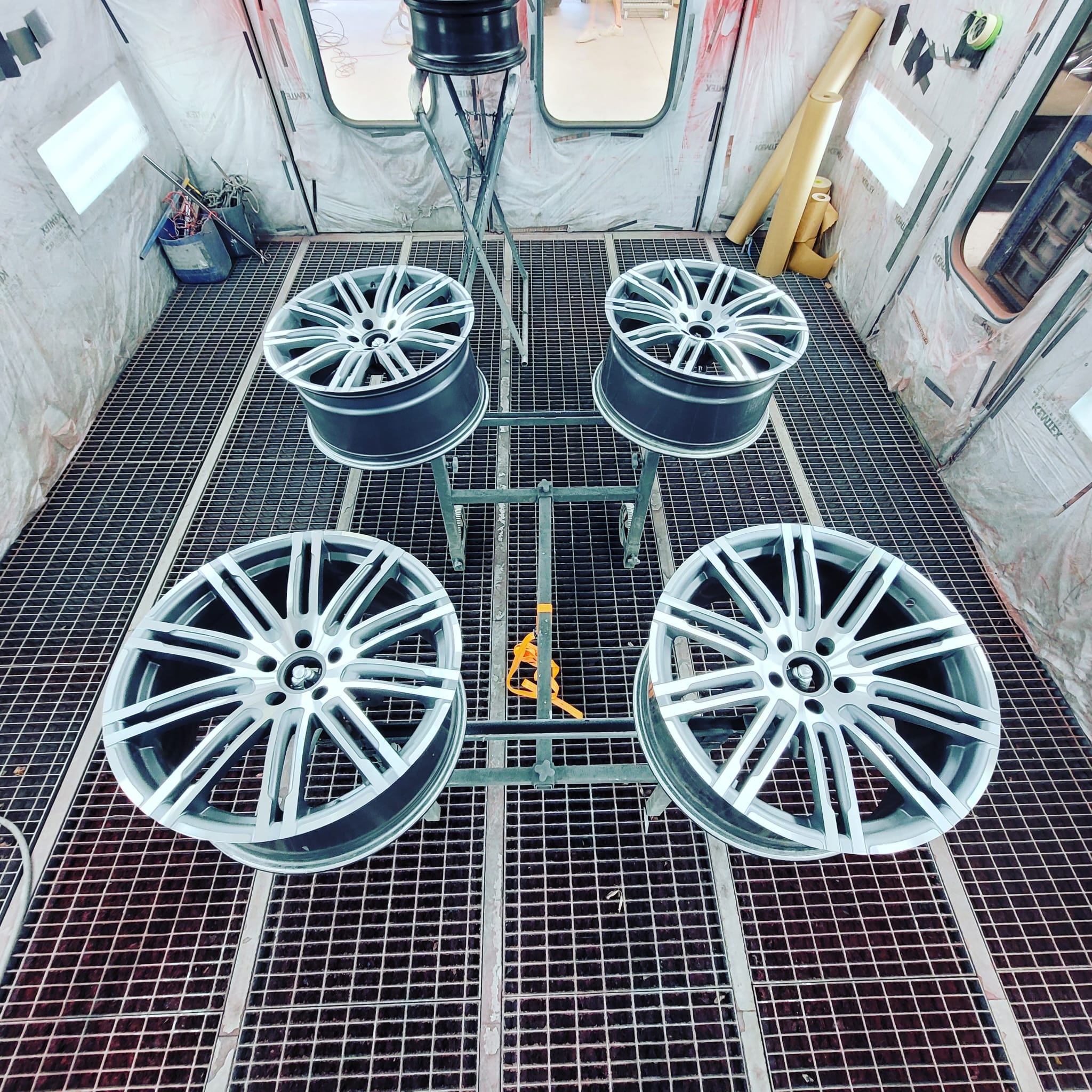 Quatre jantes de voiture en alliage disposées sur un support métallique dans une cabine de peinture industrielle.