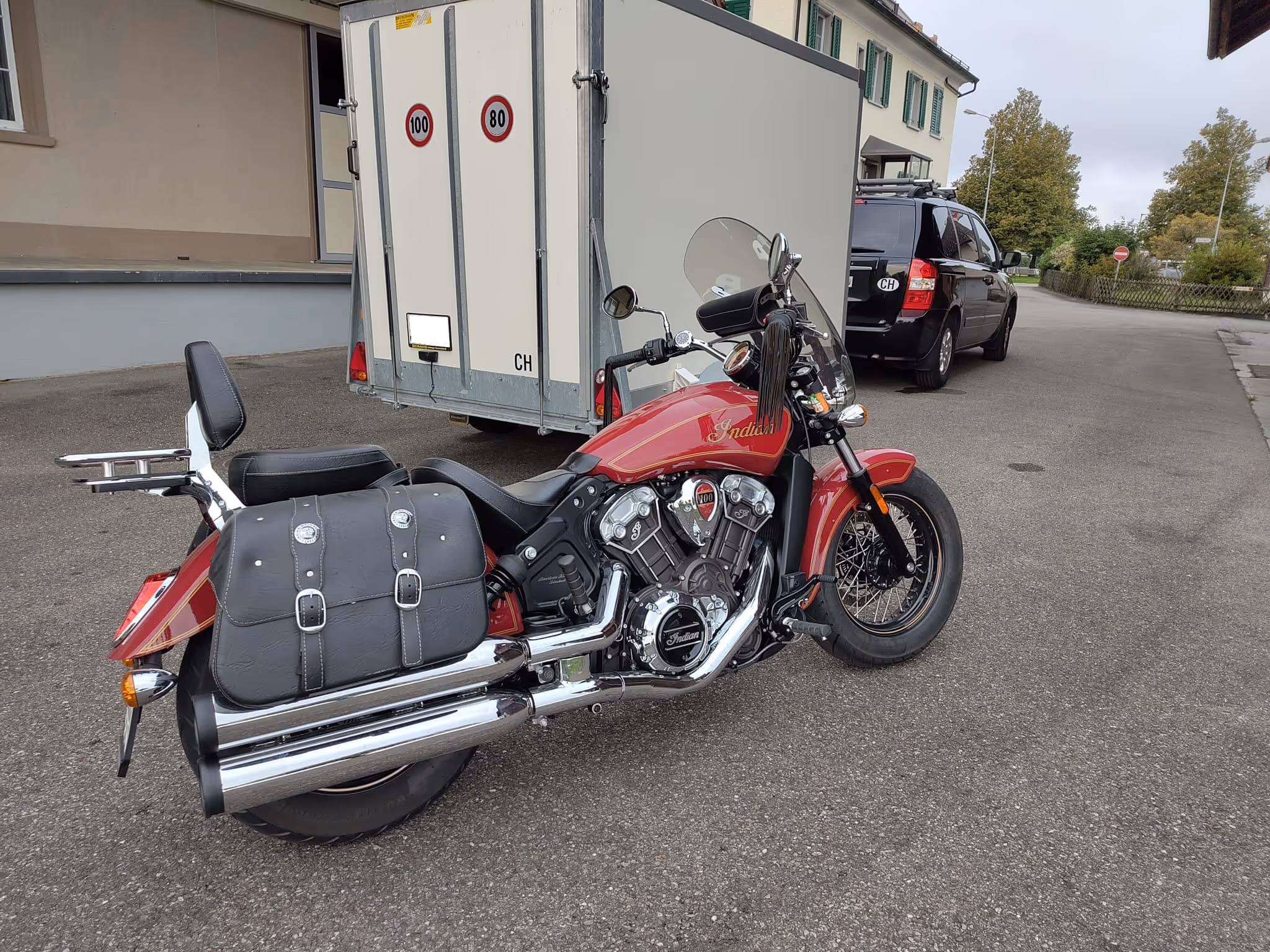 Rotes Indian-Motorrad mit schwarzer Ledertasche auf einem Asphaltparkplatz vor einem weißen Anhänger und einem schwarzen Auto.
