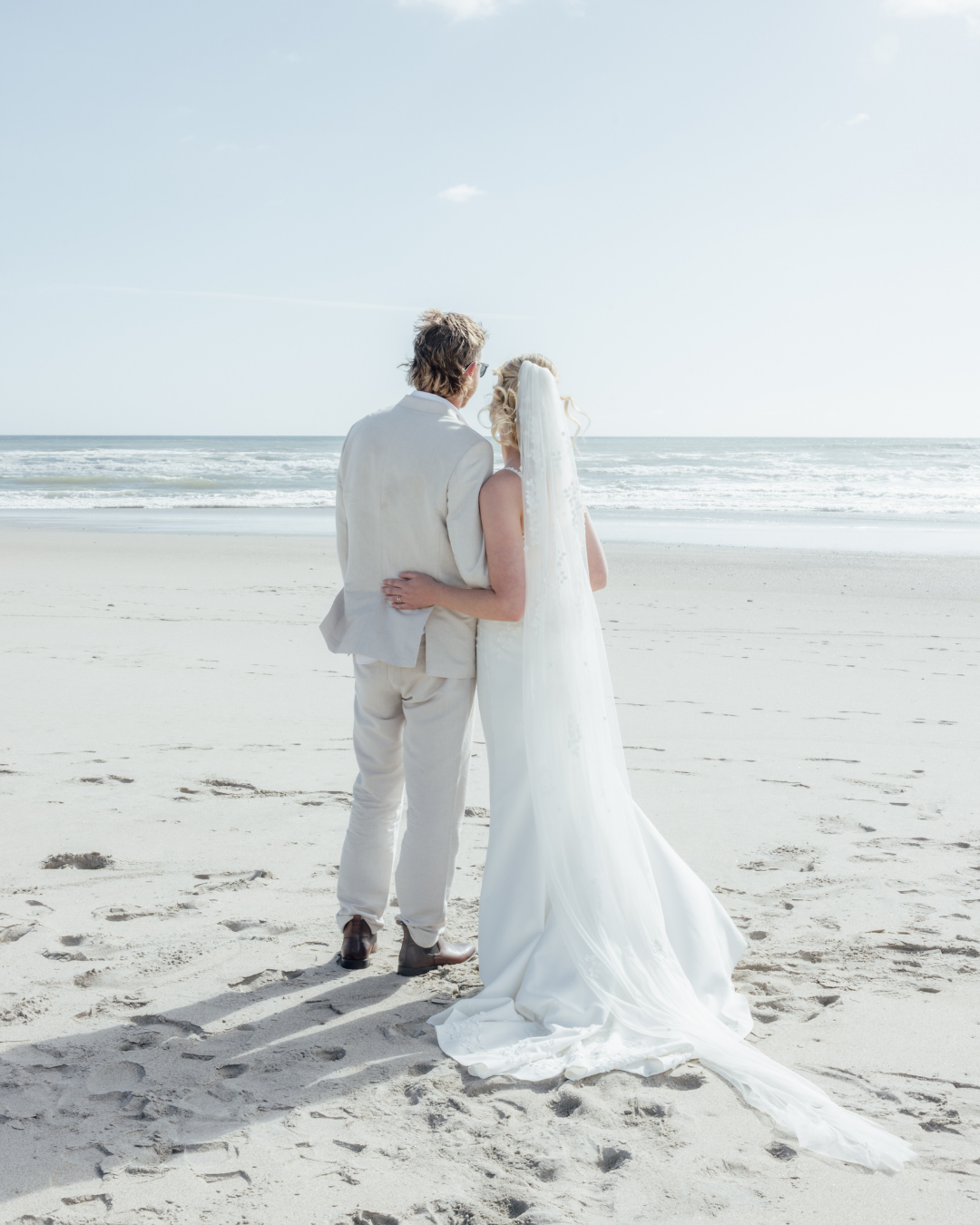 Bride and groom embracing on a sandy beach with ocean waves in the background.