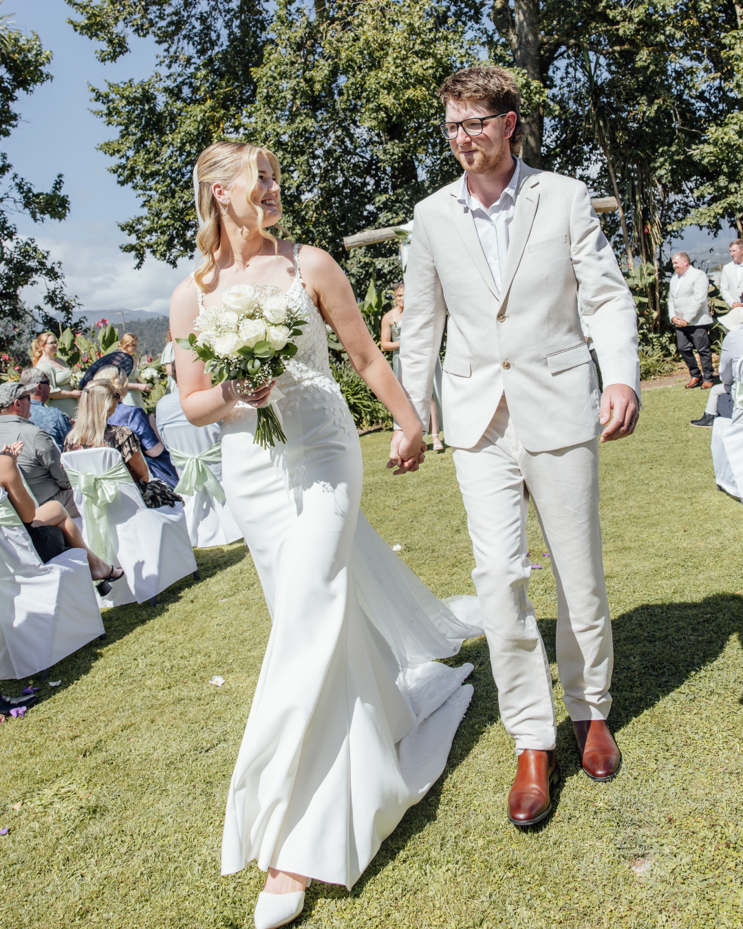 Bride in white dress holding a bouquet and groom in light suit walking hand in hand outdoors at a wedding ceremony.