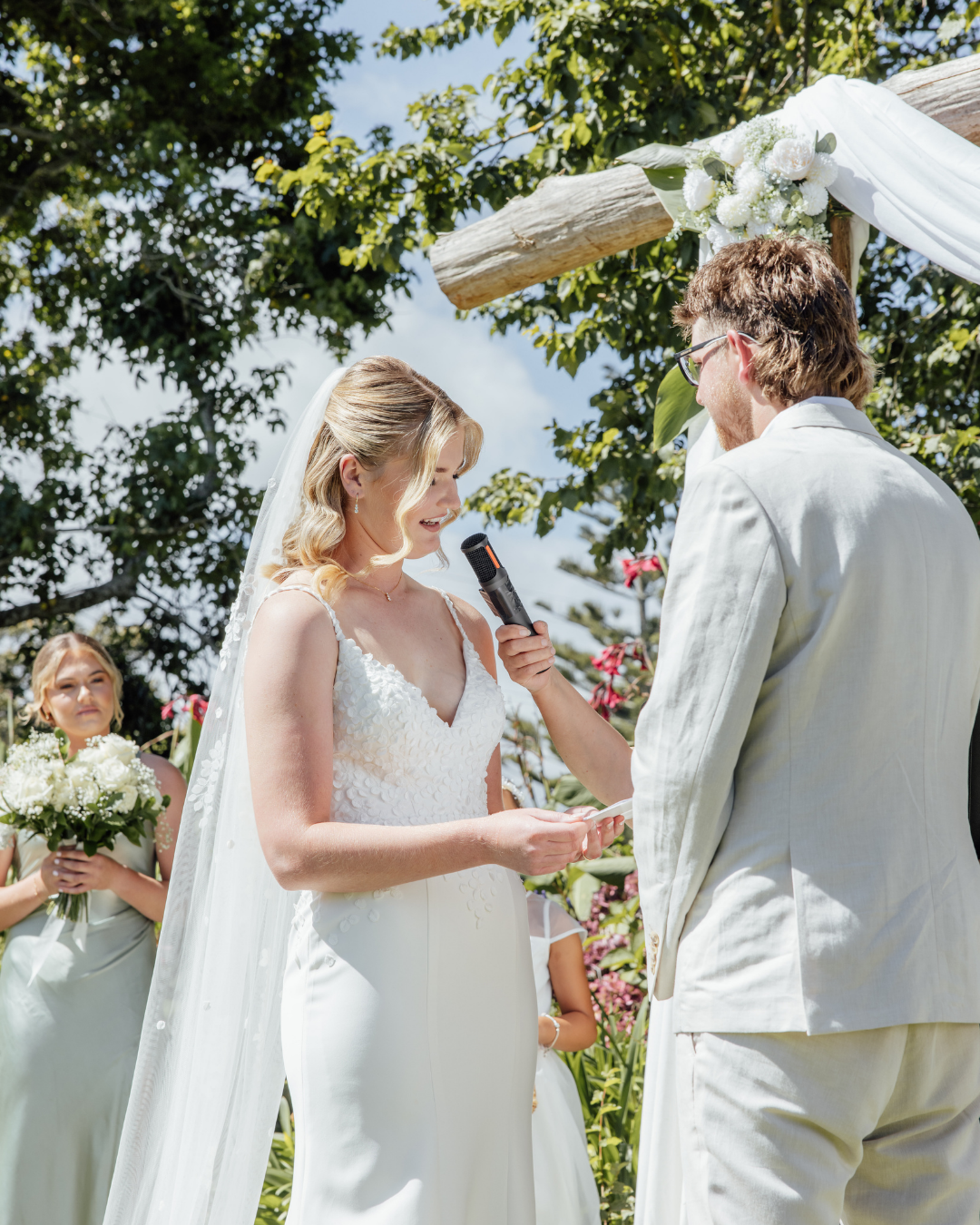 Bride in a white dress reading vows while groom in a light suit holds a microphone during an outdoor wedding ceremony.