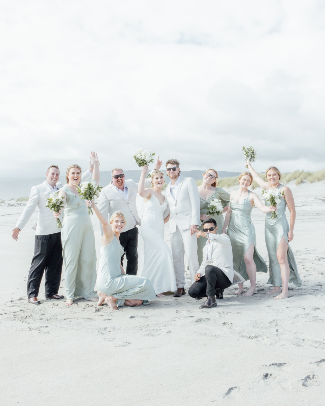 Bride and groom with bridesmaids and groomsmen cheerfully posing on a sandy beach with bouquets and light-colored attire.