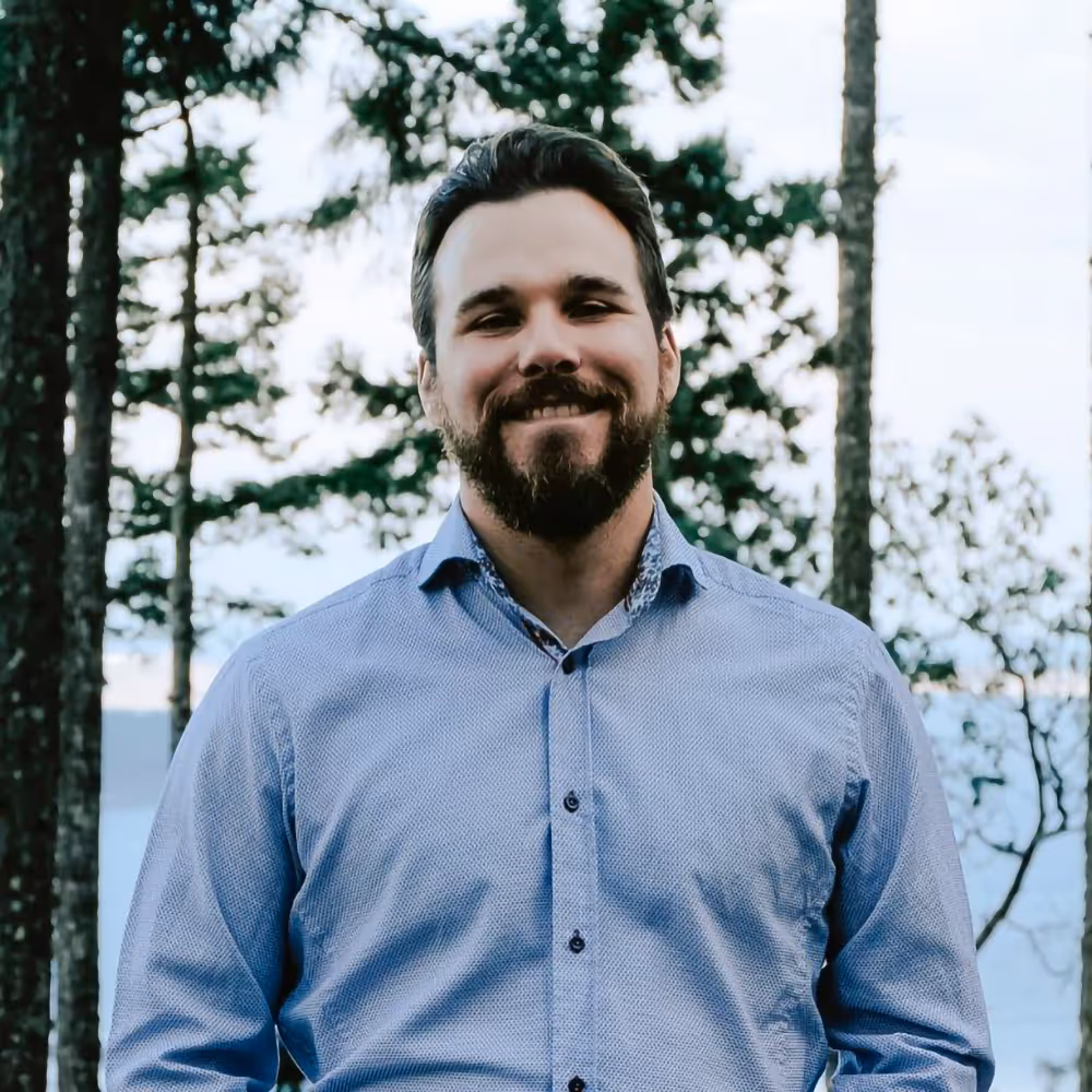 Smiling man with a beard wearing a blue button-up shirt standing outdoors with trees and a lake in the background.