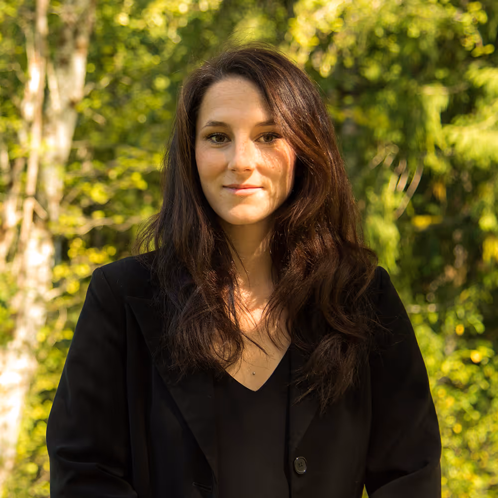 Woman with long brown hair wearing a black blazer, standing outdoors with green foliage in the background.