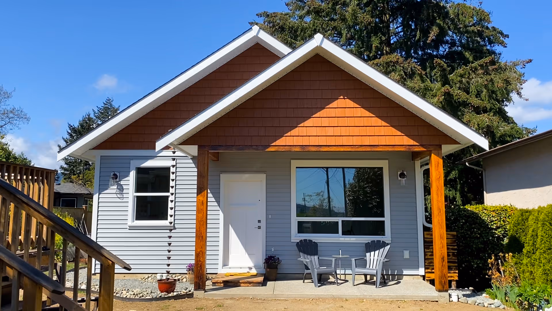 Small modern gray house with a white door, large window, and two gray chairs with a small round table on the front porch.