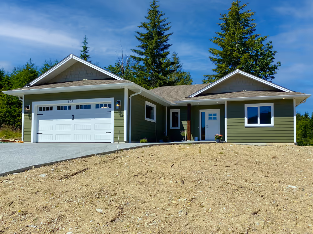 Single-story green house with a two-car white garage door, front entrance with white door, and surrounding trees under a blue sky.