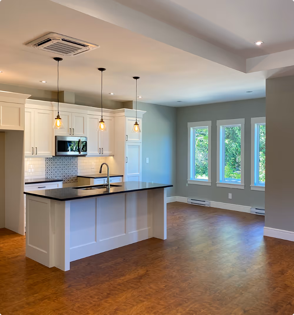 Modern kitchen with white cabinetry, black countertop island with sink, three pendant lights, and large windows showing greenery outside.
