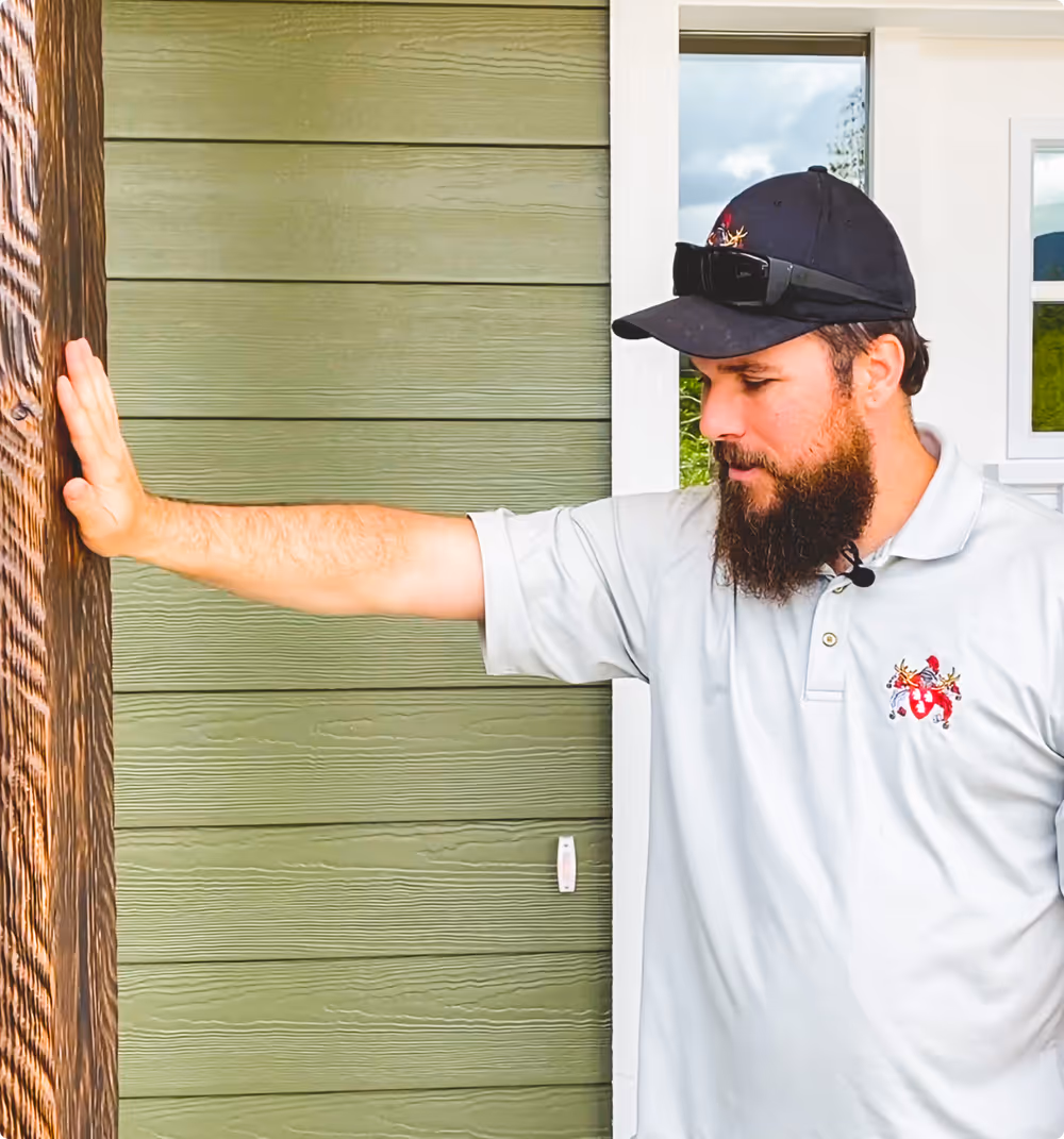 Man with a beard and black cap touching a wooden door frame on a green exterior wall.