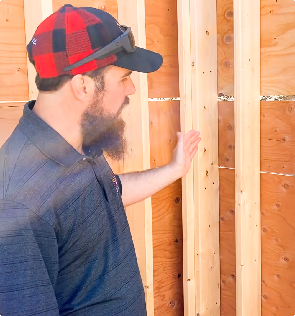 Man with a beard wearing a red and black checkered cap and dark shirt inspecting wooden framed wall studs.