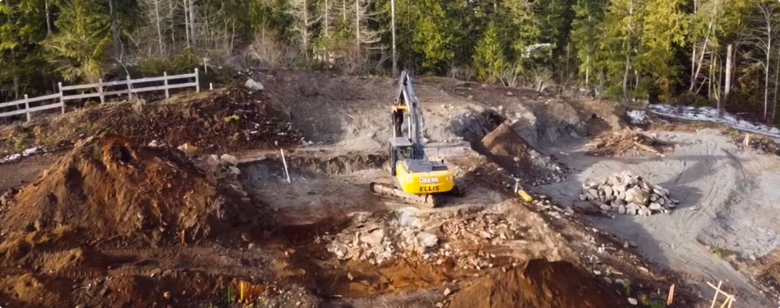 Yellow excavator digging at a construction site surrounded by dirt piles and trees in the background.
