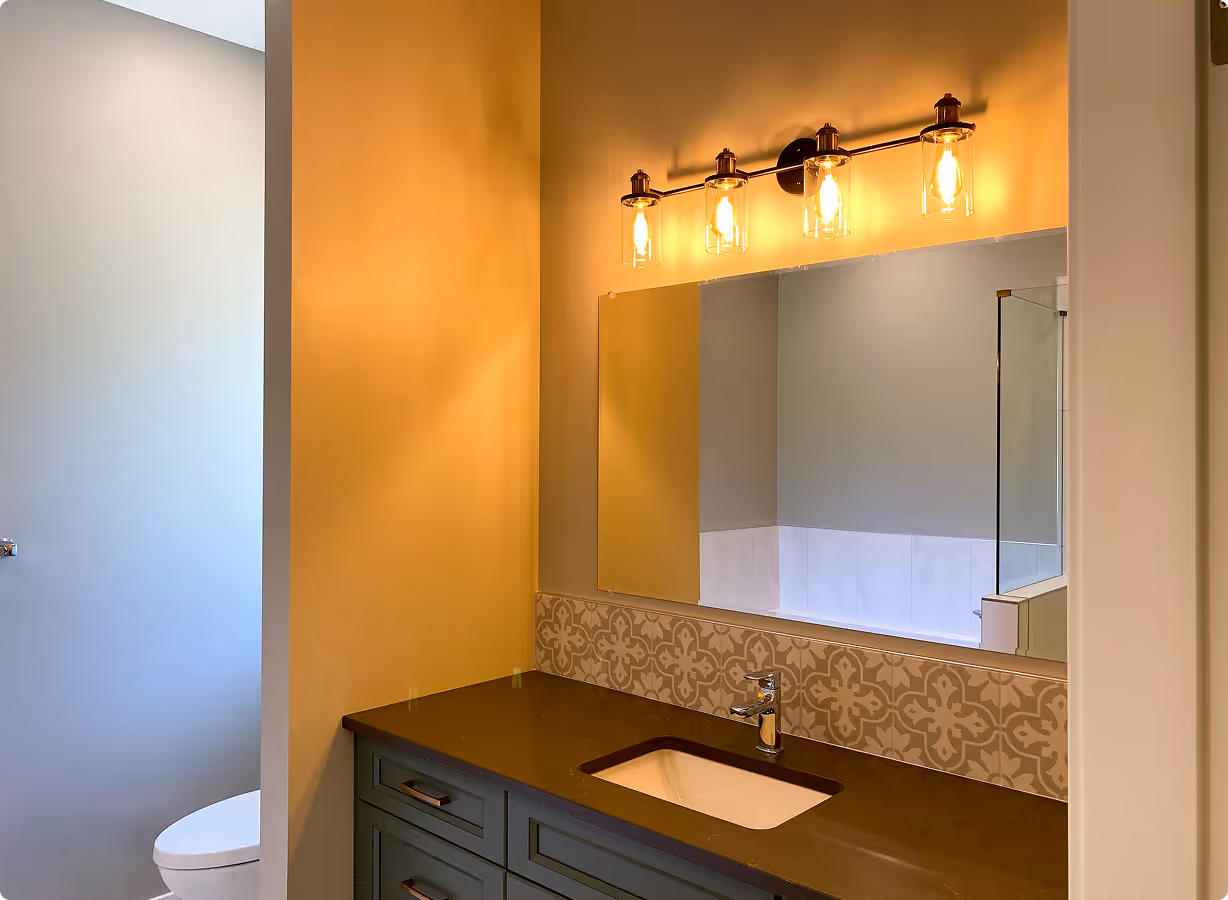 Bathroom vanity with dark countertop, rectangular sink, patterned backsplash, and three-light fixture above mirror.