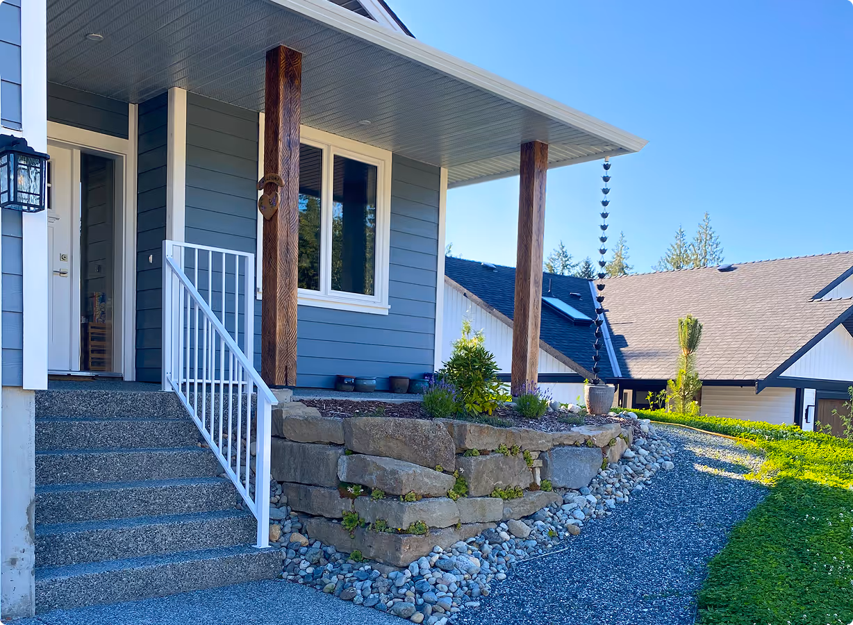 Front porch of a house with gray stone steps, white railing, wooden columns, stone retaining wall, and a gravel walkway curving to the right.